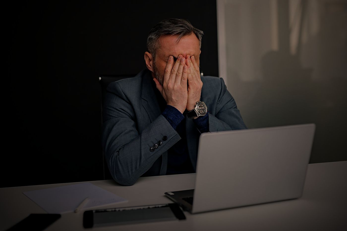 Homem grisalho de terno, à noite na mesa de trabalho, com as mãos no rosto